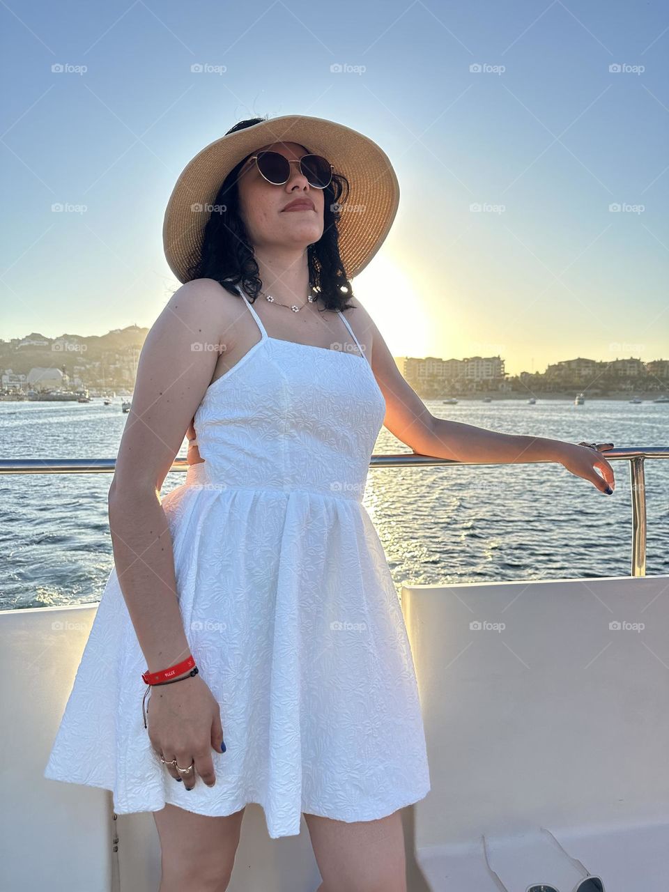 Woman wearing a hat and sunglasses, standing on a boat in the ocean, with the sunset at the background.