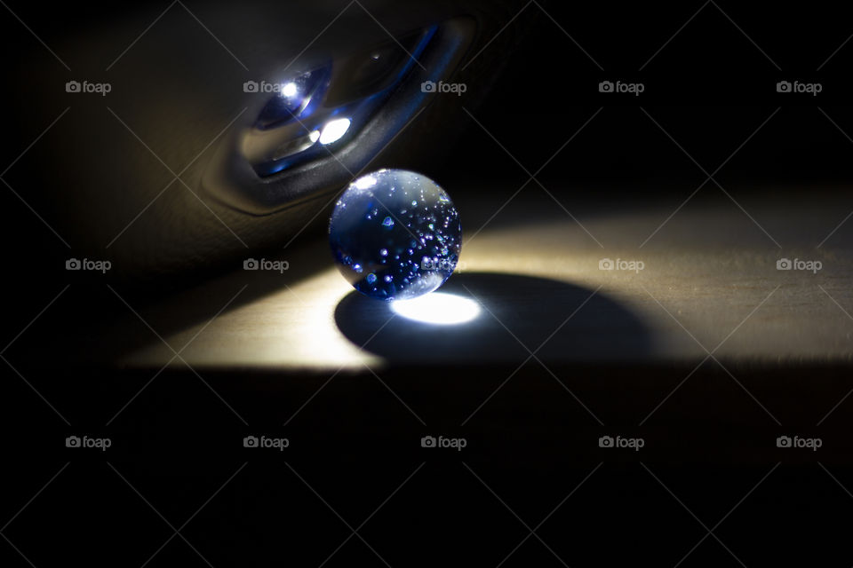 A portrait of a blue colored marble on a wooden surface lit by the flashlight of a smartphone.