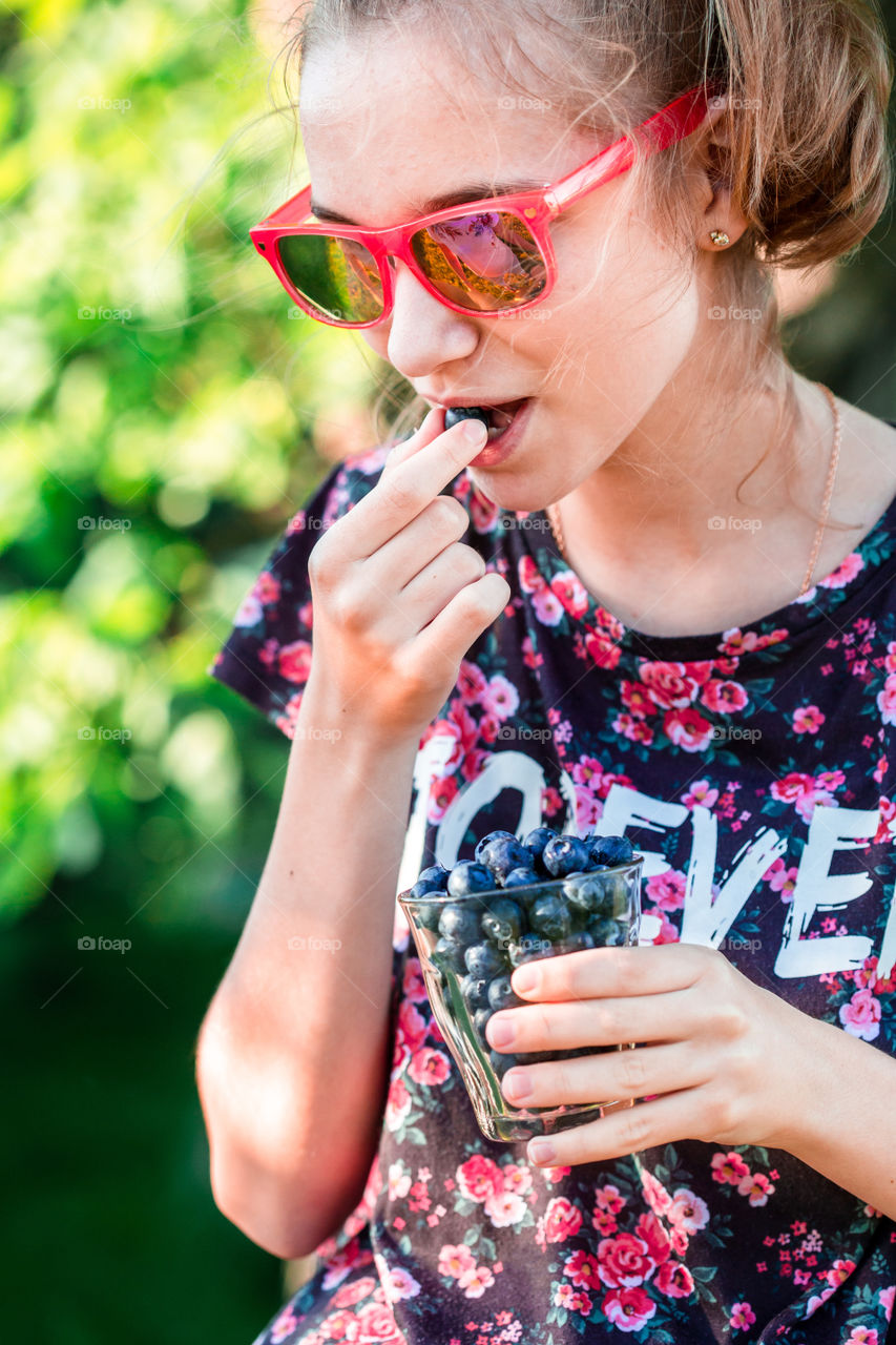 Happy girl enjoying the fresh blueberries outdoors. Teenager girl wearing flowery red-blue blouse and red sunglasses. Garden in the background