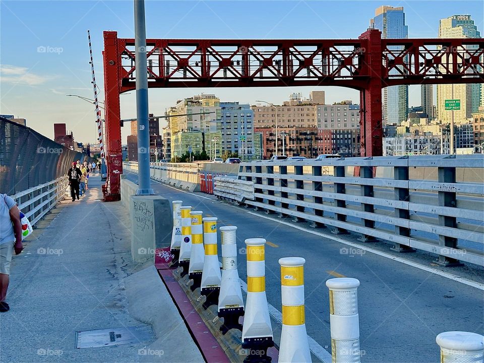 This is the pedestrian and bicycle lane of the “Pulaski Bridge” at “Newtown Creek” connecting “Greenpoint”, Bklyn to LIC, Queens in the direction we are headed. The ornamented red metal gate reminds of the “Bauhaus” style. 2024. Hypnotic Productions