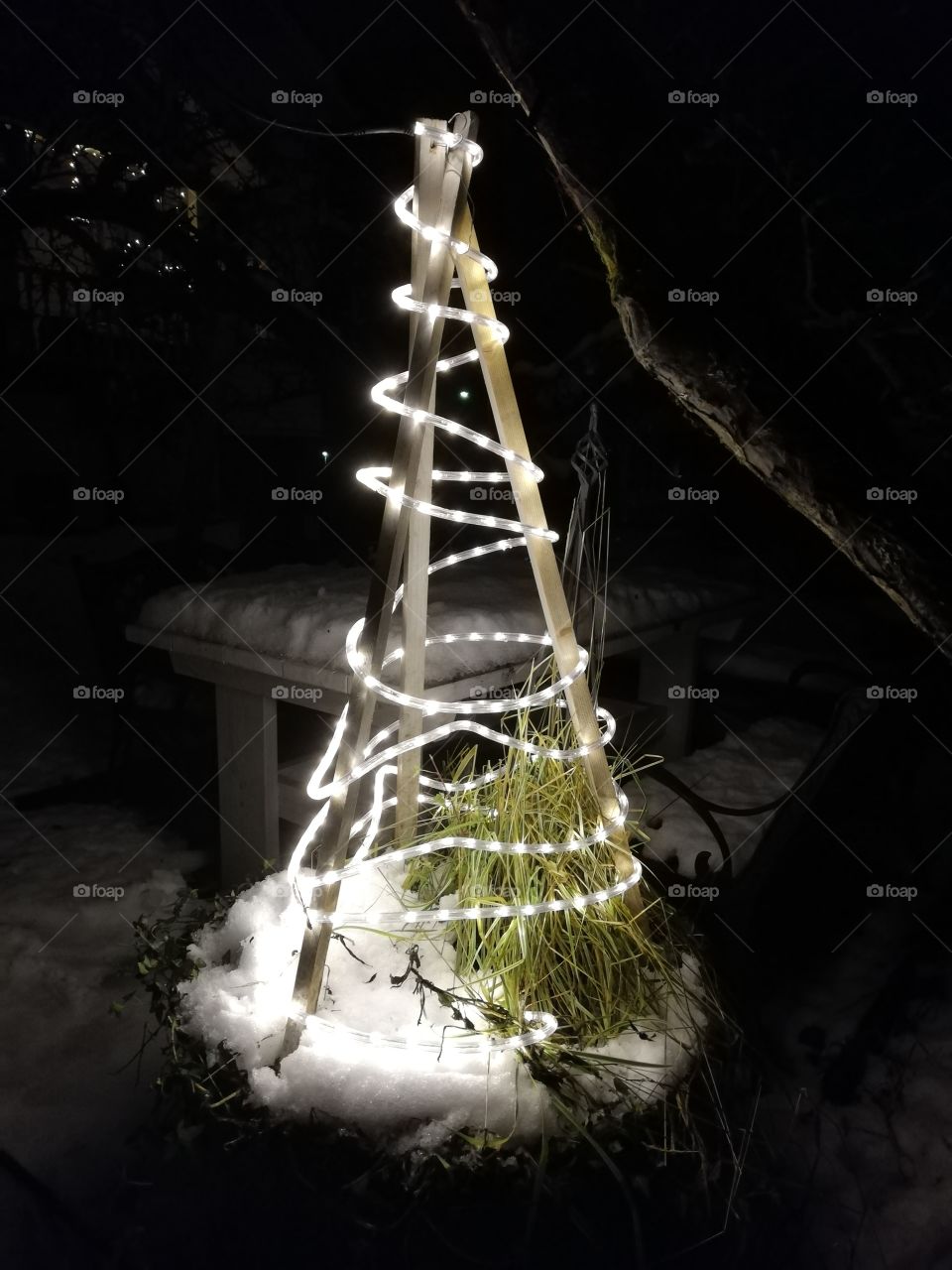 A Christmas tree made of unpainted slats around green grass and snow. The bright lights in a plastic cable weaving the skeleton.