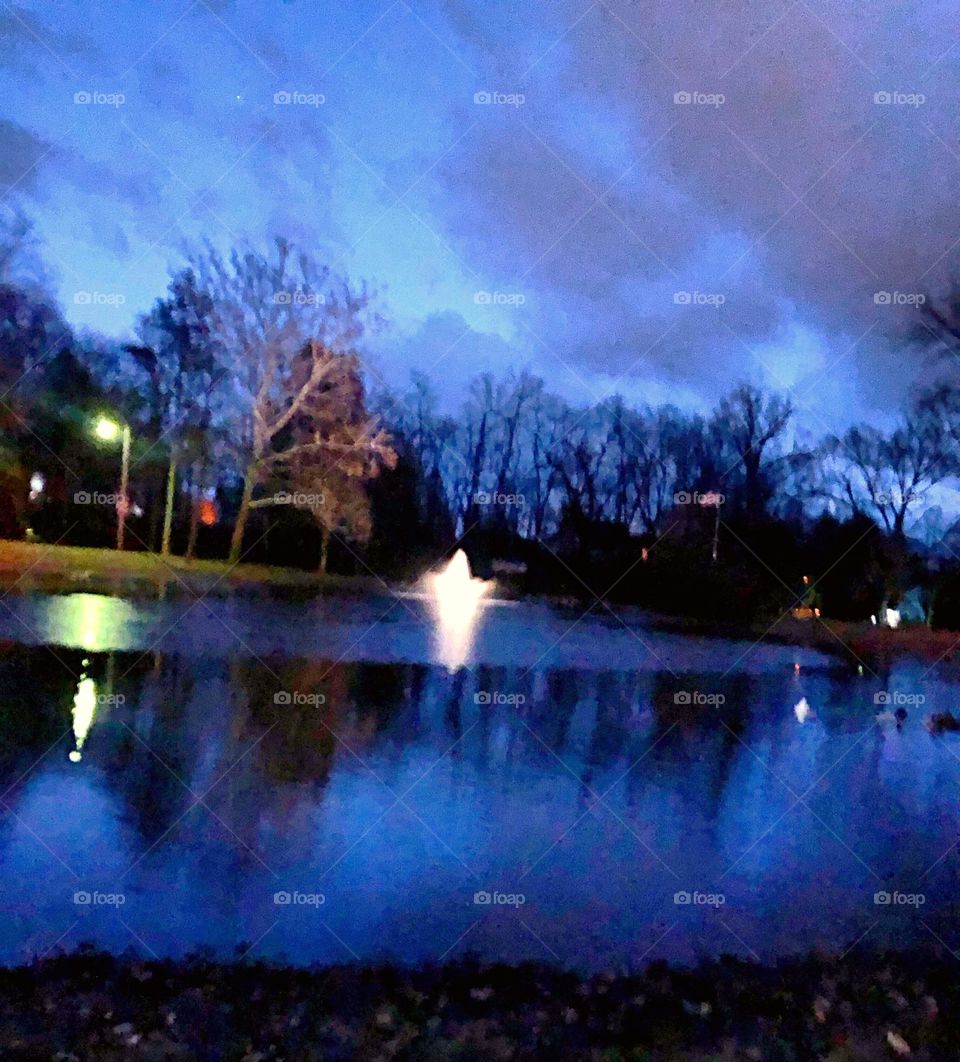 A almost nighttime photo of a pond and it’s reflection in a park surrounded by trees and lit by street light and fountain light with clouds overhead 