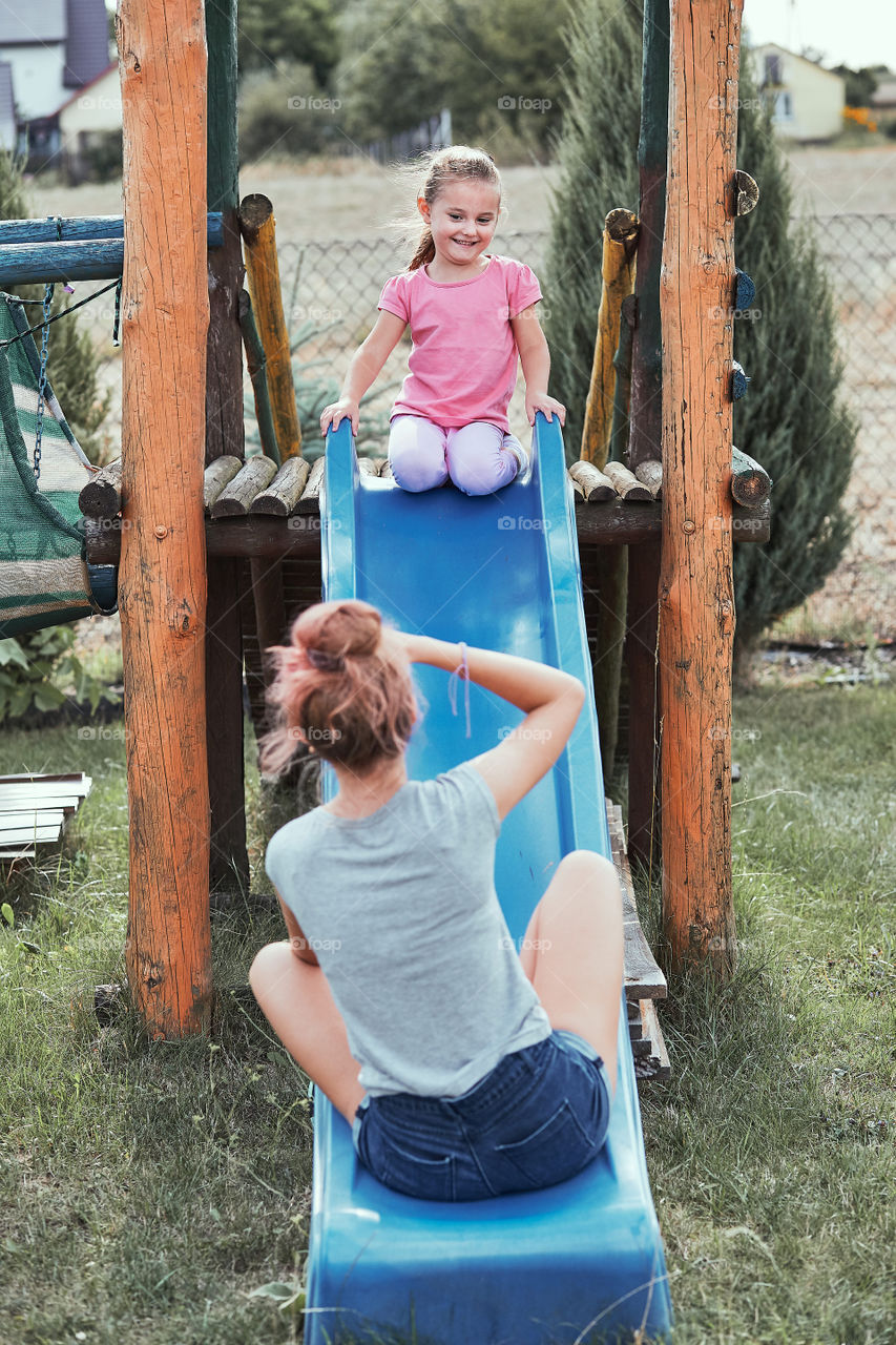 Teenage girl taking a photo her younger sister in a home playground in a backyard. Happy smiling sisters having fun on a slide together on summer day. Real people, authentic situations