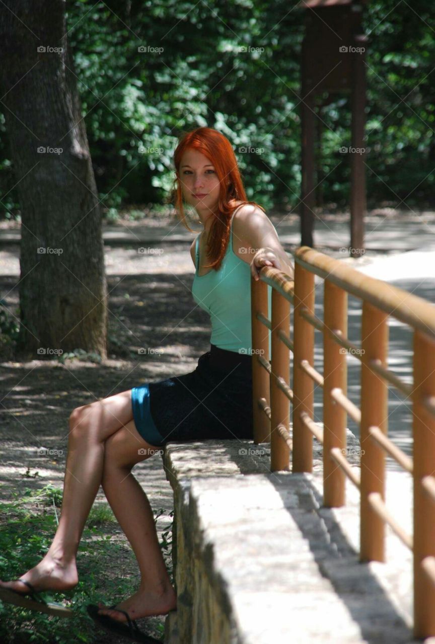 redhead woman sitting against railing of bridge in woods