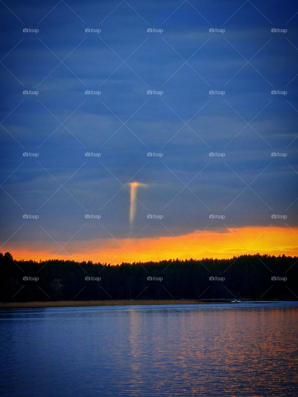 Sunset on the lake. Trees on the shore of the lake. The whole sky is covered with dark clouds. Only above the trees is a yellow sunset. A ray of light breaks through dense clouds.