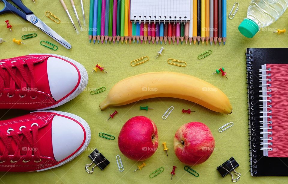 Let's go to school. Set for school with a snack on the background of green paper. Sneakers, banana, apples, paper clipsbuttons, colored pencils, notepad, copybooks, paint brushes