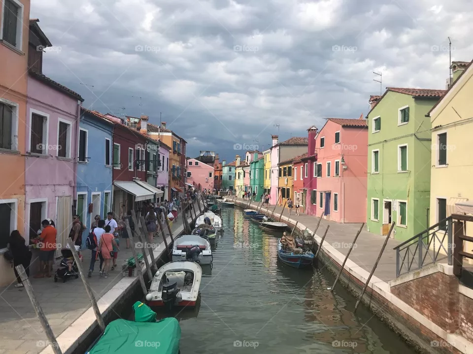 Explore the colorful world,small canal with a few canoes anchored, 2 sides are colorful houses, colors are reflected into the water. A lot of gray clouds in the sky, it's going to rain , there are many strong winds. many people