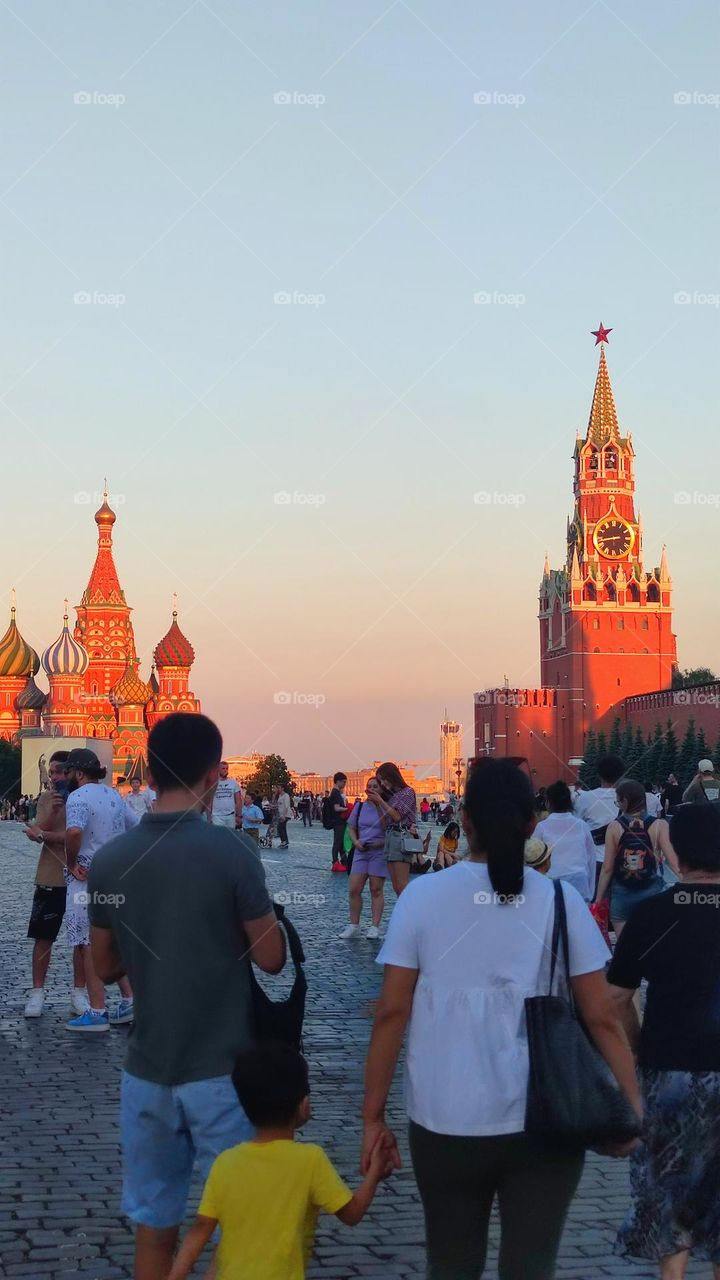 Summer evening. Red Square where people walk. Basil's Cathedral and the Spasskaya Tower with chimes in the orange rays of the setting sun. Moscow