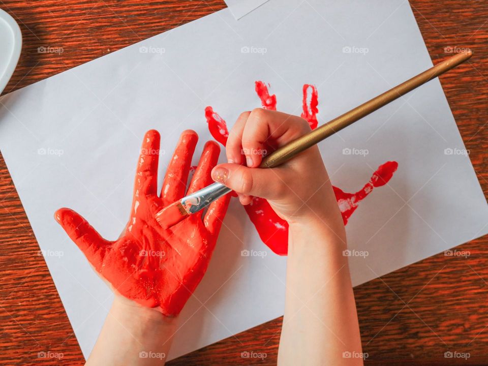 Little caucasian girl paints her hands with koas paint with a brush and prints them on a white sheet of paper, sitting at a wooden table, close-up top view. Kids creativity concept.