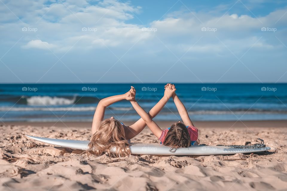 two little girls are lying on a surfing board on the beach and holding each other hands