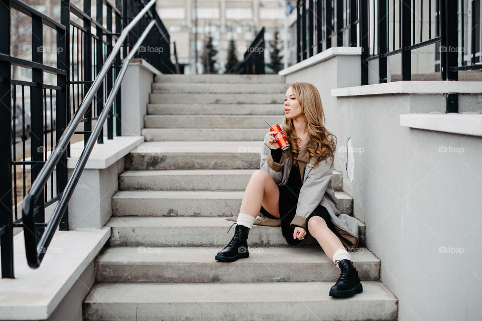 A woman in grey coat sitting on stairs and drinking Coca-Cola