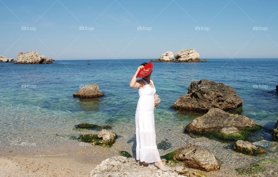 A woman in white dress with a red fan on the seashore