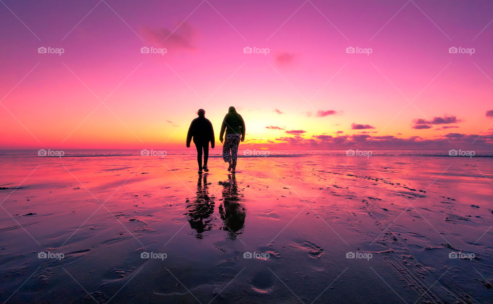 Silhouette of two people walking on the beach towards a colorful dramatic sunset which is reflected on the beach