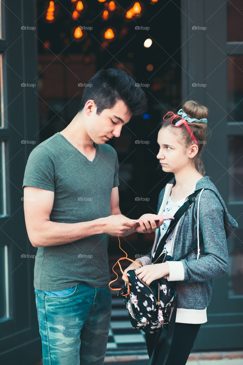 Couple of friends, teenage girl and boy, using smartphones, talking together, standing on street in center of town, spending time together