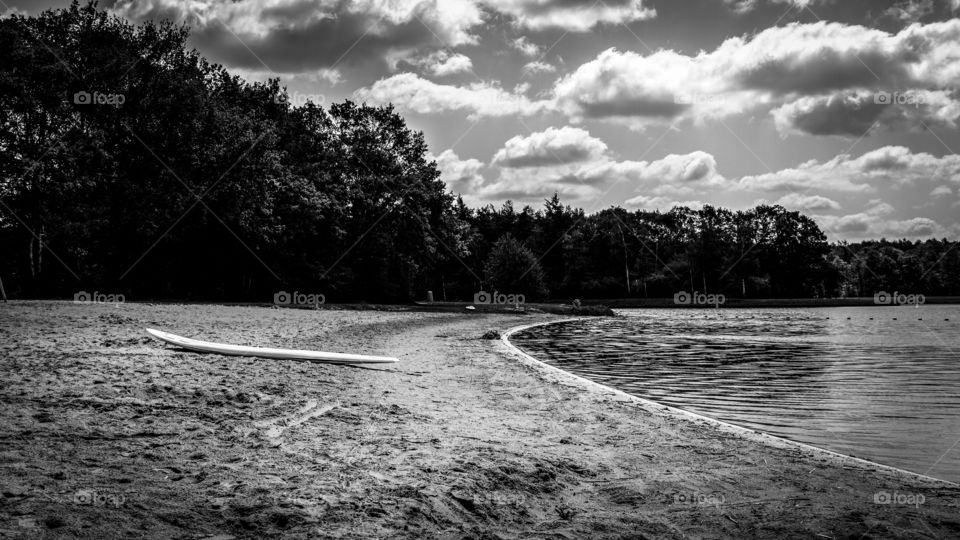 A black and white landscape picture of a beach at a lake in Bussloo, Holland. the beach and the lake are surrounded by a forest. on the beach there is a surfboard.