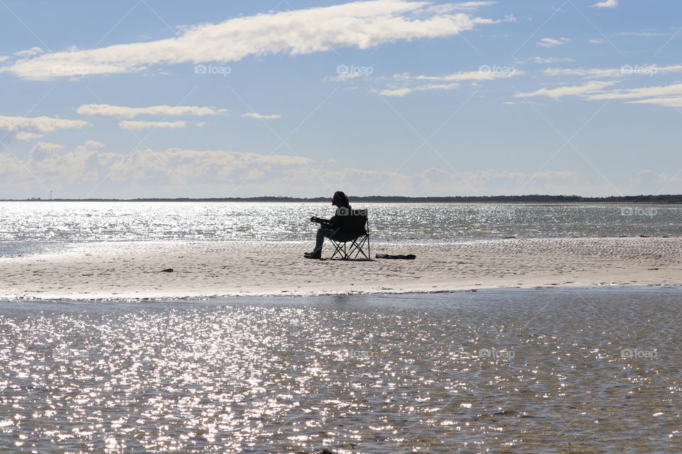 Stranded, lone guitarist singer sitting in chair at beach as tide rises silhouette 