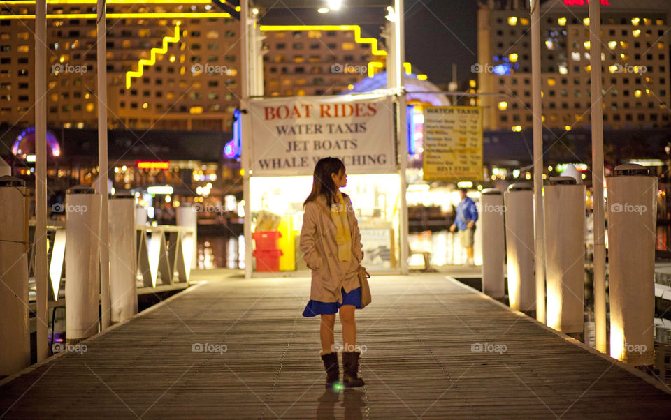 Lady in docking bay looking away for boat at night, city lights