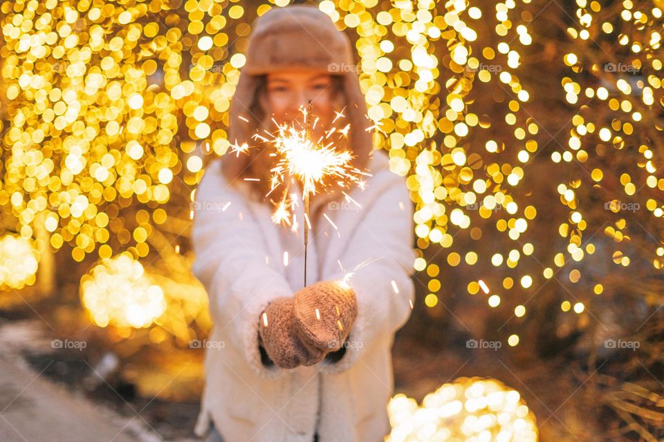 Young happy woman with curly hair in fur jacket having fun with sparkler in winter street decorated with lights