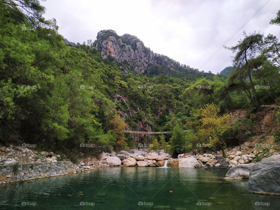 A beautiful nature photo with a lake, a micro waterfall and rocks in front, trees and a mountain in a cloudy sky.