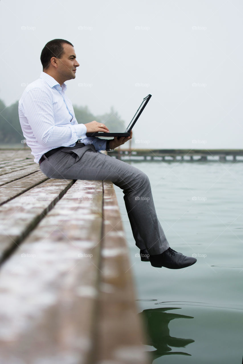young man with laptop with smile, sitting on a lake dock