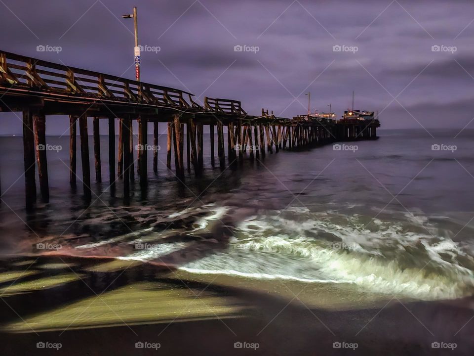 Looking down the beach at Capitola by the Sea in California at night with a soft cloudy sky, the wharf jutting out into the ocean that sustained damage in the January 2023 storms that ravaged the coastal region