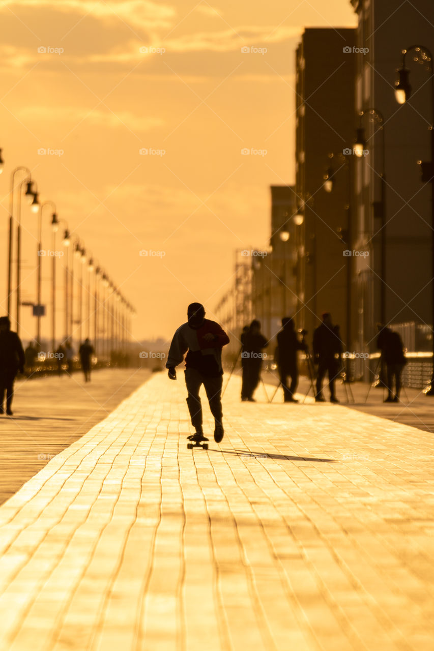 Silhouette of a skateboarder riding down a boardwalk, as the sun sets and casts beautiful golden light and shadows on the wooden path