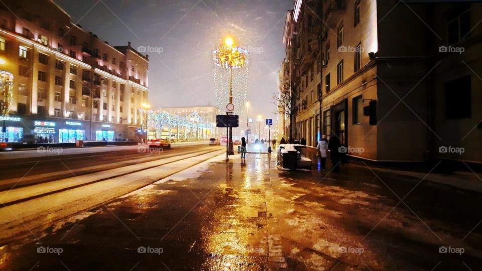 A wide avenue on a winter evening, sleet, inclement weather, wet asphalt, reflections of yellow light from street lamps on a wet sidewalk