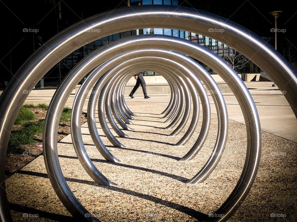 A spiral, metal bike rack with a view from one end to the other and a persons legs walking through the opposite end- abstract view