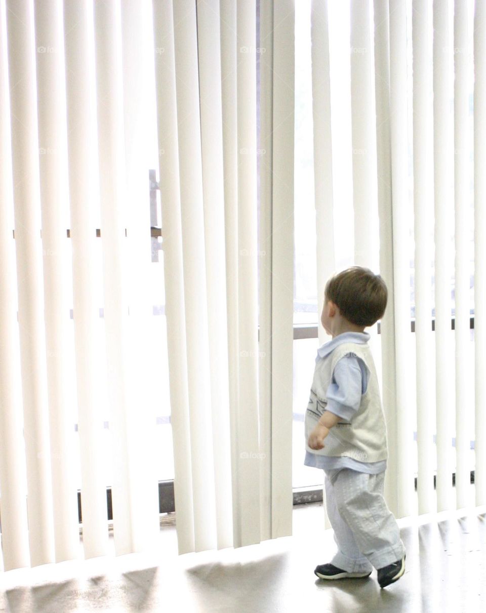 Little boy looking through white blinds