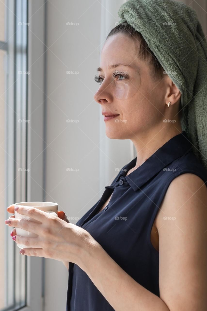 A young girl after a shower stands at home near the window drinking tea
