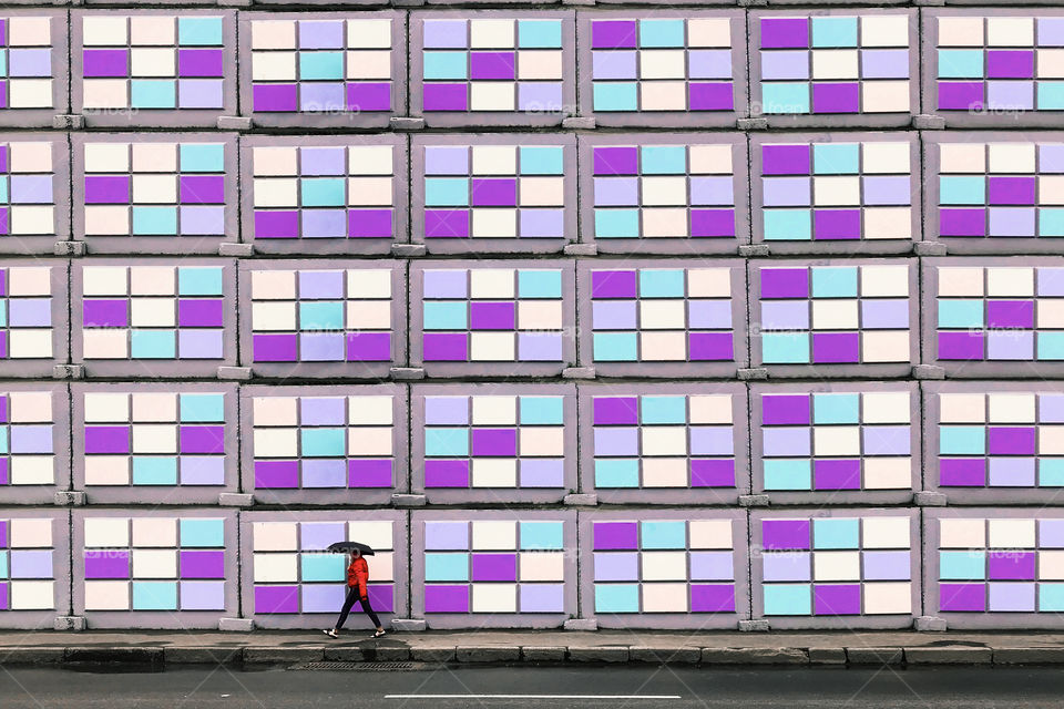 Tiny human walking by the colorful purple wall with umbrella 