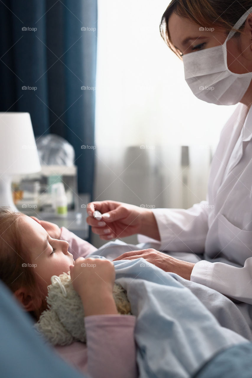 Doctor visiting little patient at home. Measuring the temperature of sick girl lying in bed. Woman wearing uniform and face mask. Medical treatment