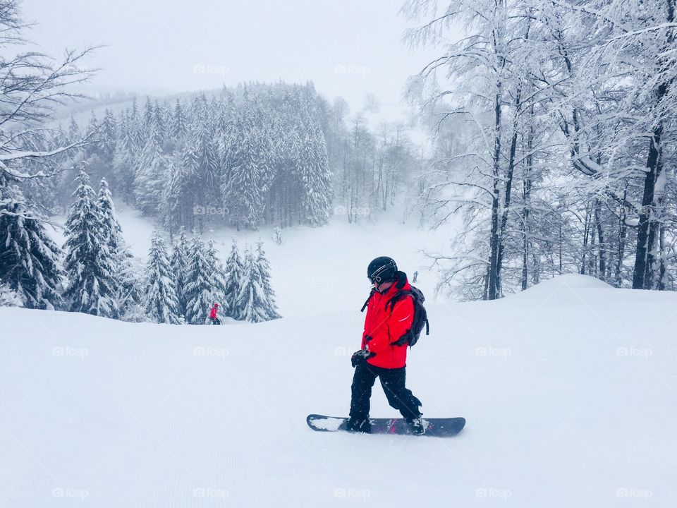 Man in red jacket with snowboard going down the slope surrounded by trees covered in snow 
