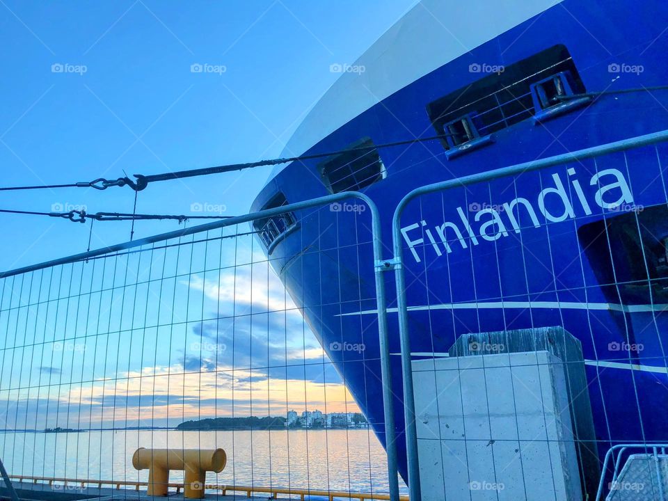 Close view of the bow of the blue ship Finlandia behind a lattice fence close to the pier with smooth sea water surface and clear transparent sky with sunlighted clouds on the background