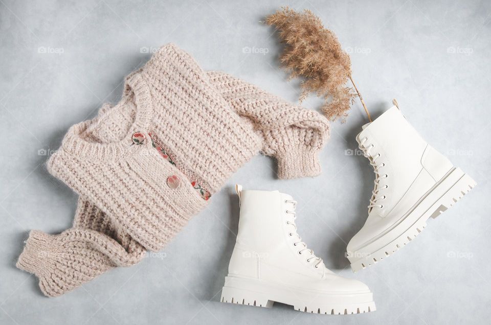 Beautiful white boots on a rough tractor sole with eco leather lacing with a knitted beige cardigan and a bouquet of dried flowers lie on a light cement background,flats Lay. The concept of fashionable women's clothing and shoes.