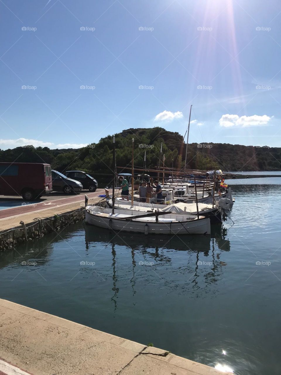Small boats moored in quaint menorcan harbour. Shadows of the boats are vibrant in the still water. Lovely blue sky and green hills in background 