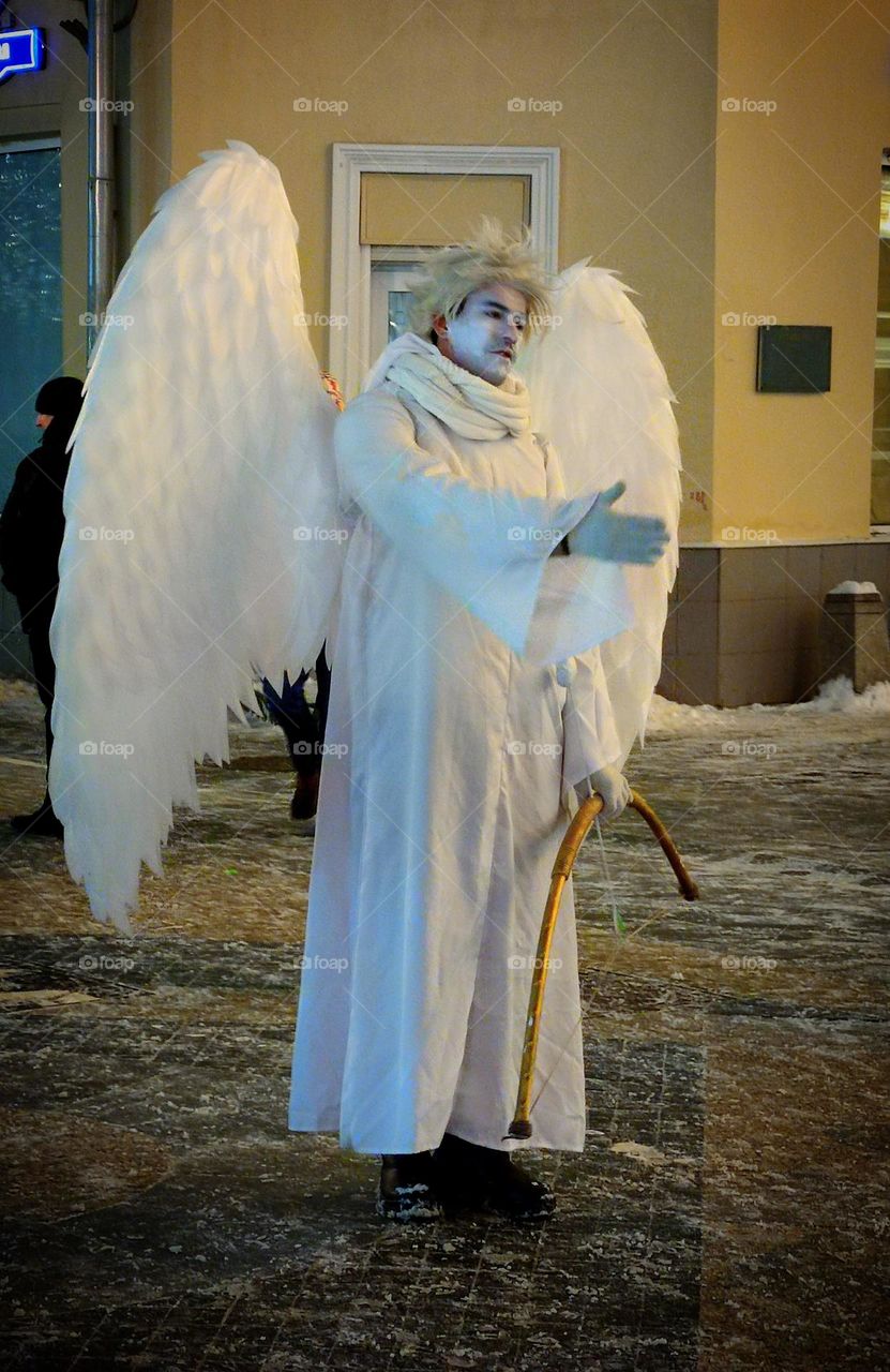 Sadness. A man dressed as a white angel is standing on the street. The angel holds out one hand to people. The face and posture of the angel shows the emotion of sadness