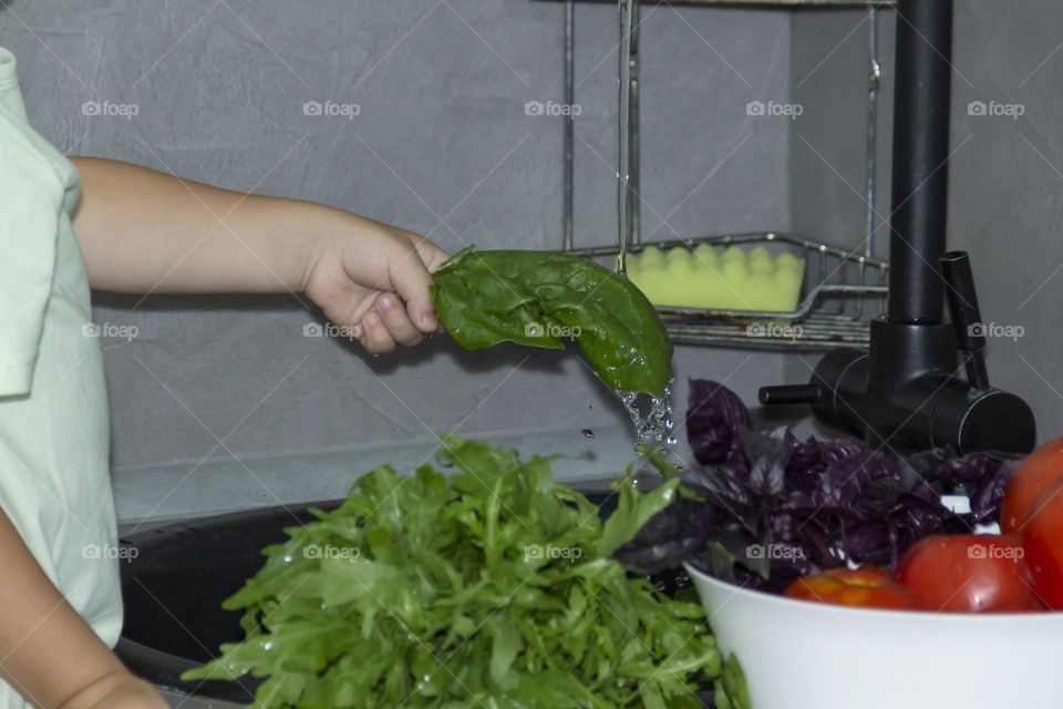 A small child washes fresh, green, vegetarian vegetables under the tap in a black sink to prepare salads and other dishes in a homemade gray and green kitchen.