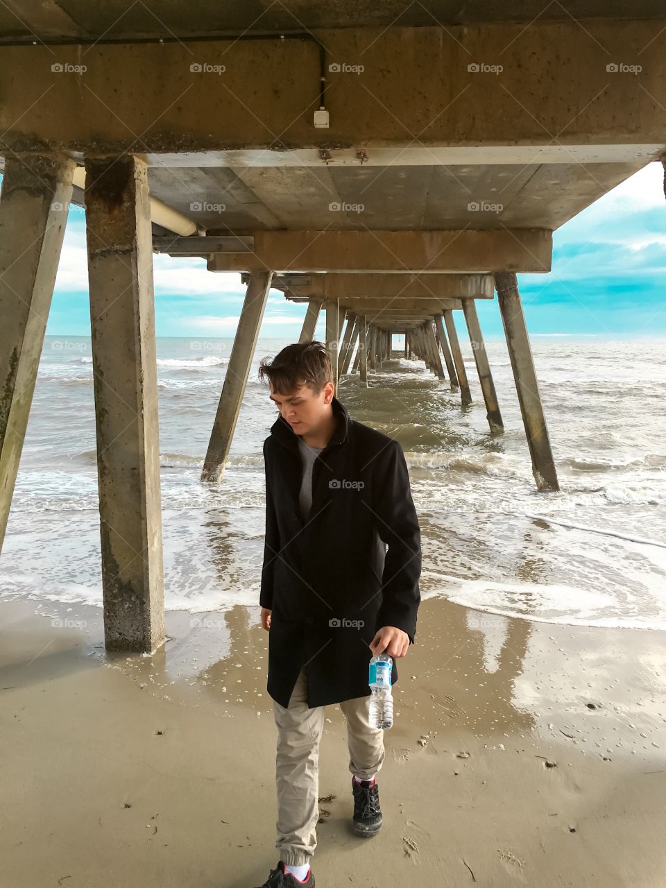 Young man in black coat standing walking underneath wood jetty at ocean