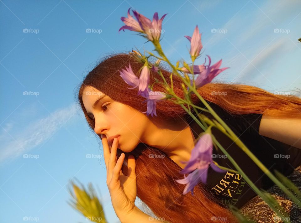 It is a photo of a young woman in the field. She has red (ginger) hair. She has brown eyes. Her face is partly coverd with Campanula patula or spreading bellflower