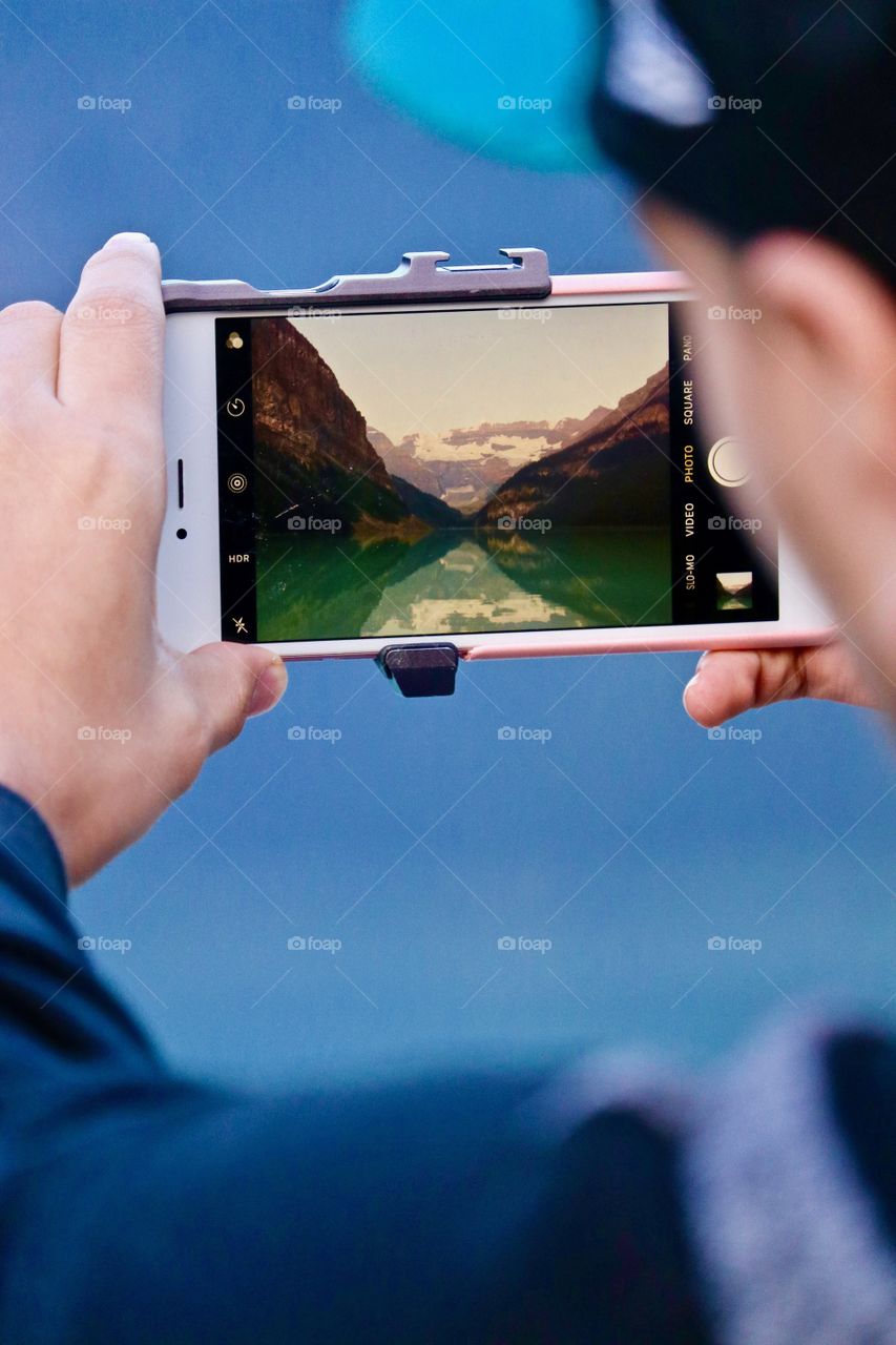 Young man photographing scenic Glacial lake and mountains with mobile phone; image captured on his smartphone; photo within a photo (Lake Louise, Banff Alberta )