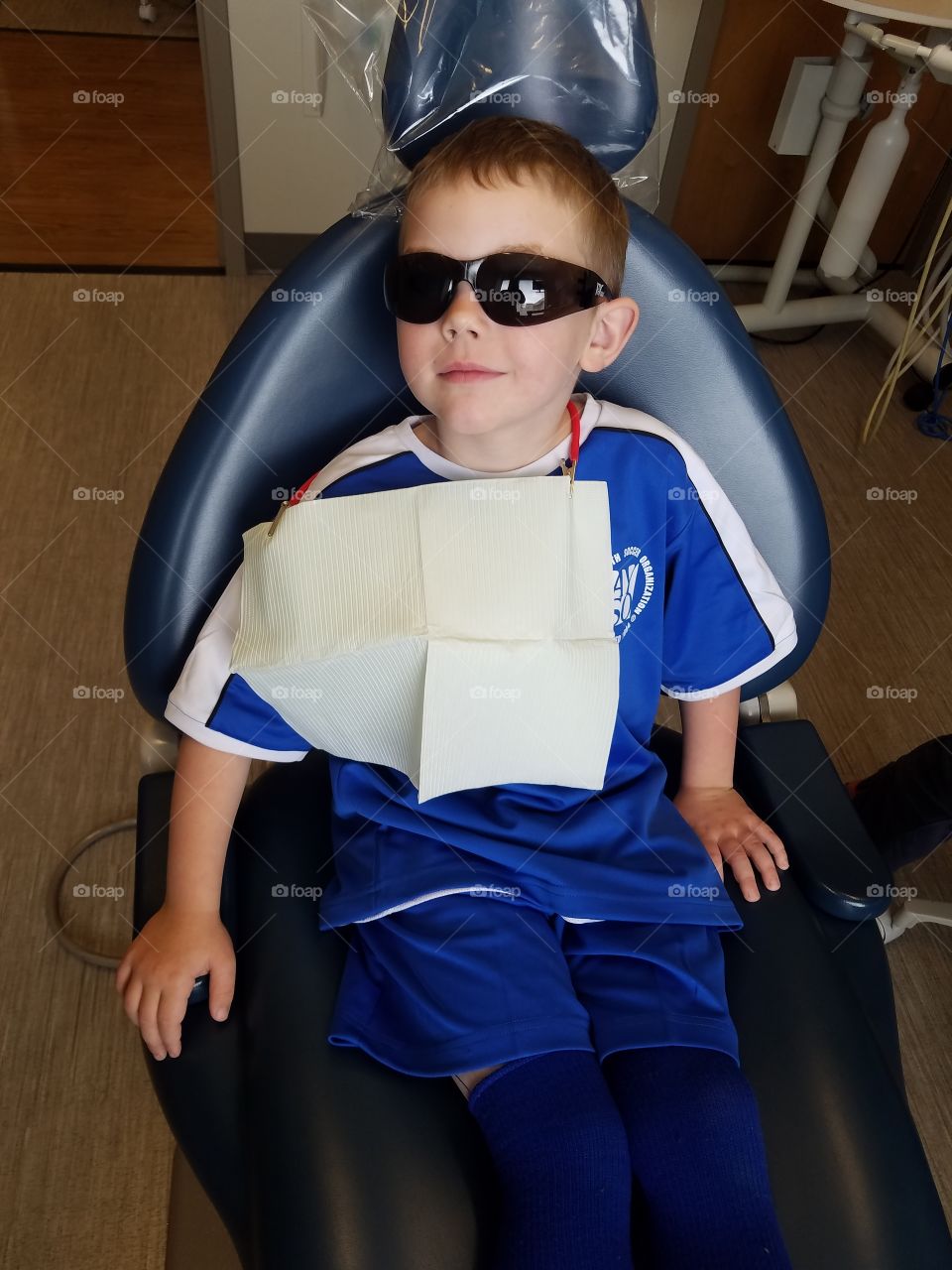 Blonde boy, in blue soccer uniform and dark glasses, sits smiling in dentist chair.