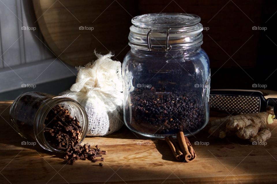 Ingredients for elderberry syrup—cloves in small glass jar, elderberries and chopped ginger in cheesecloth, dried elderberries in sealed glass jar, cinnamon sticks and ginger root with grater on wooden surface