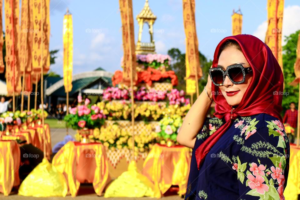 Portrait of a beautiful mother wearing a red hijab, wearing a floral patterned robe and wearing black glasses, taking a photo with a happy expression.