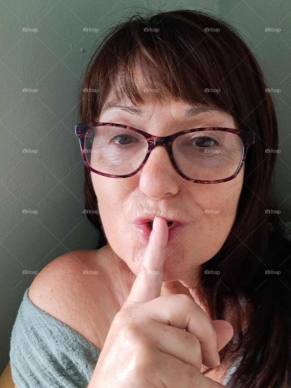 clean skin, no makeup. lady with glasses, signs of aging skin. shoulder. saying ssshh wirh finger on mouth