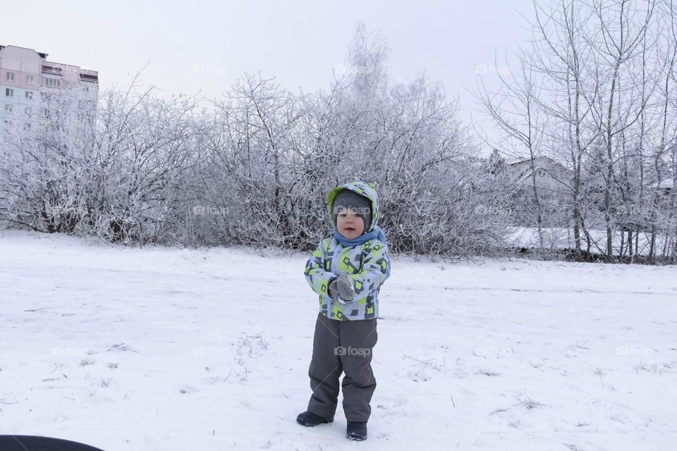 A small, carefree boy walks in winter through the white snow in the park, near the trees in the snow.