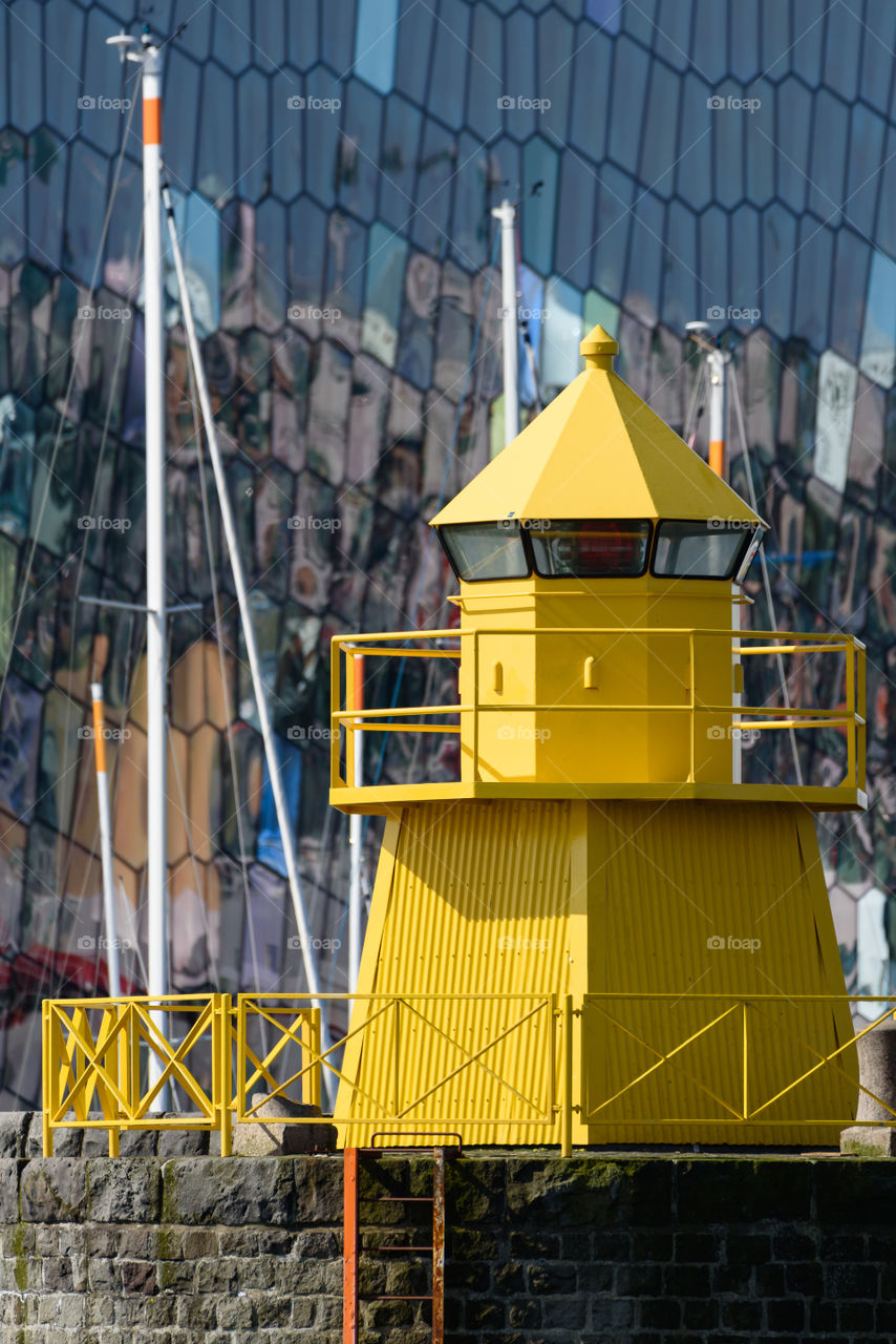 REYKJAVIK, ICELAND - JUNE 3, 2017: Yellow lighthouse in the Reykjavik, Iceland harbor with colorful and abstract glass exterior of the Harpa concert on the background with reflections in the windows and masts of the sailing boats in between.