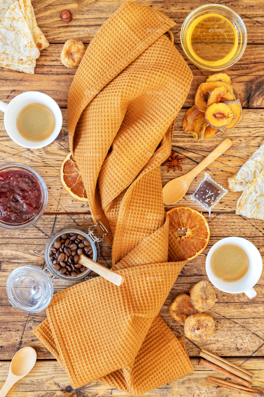 Breakfast food composition with a folded kitchen towel around which pancakes, fruit chips, nuts, honey and two cups of coffee. Healthy snack and eco friendly dining accessories. Veggie coffee time