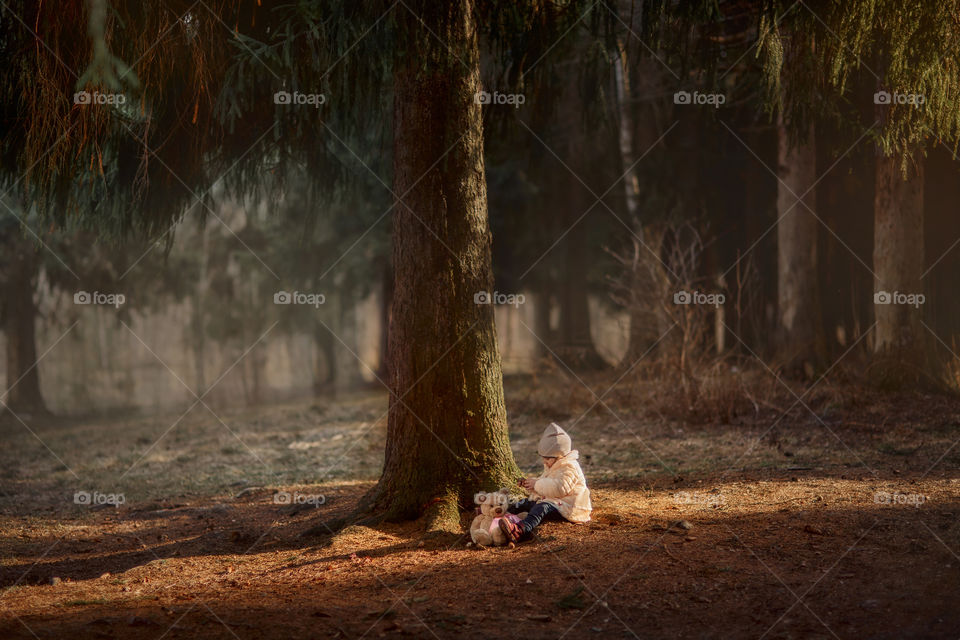 Little girl sitting with teddy bear under big spruce tree in spring park