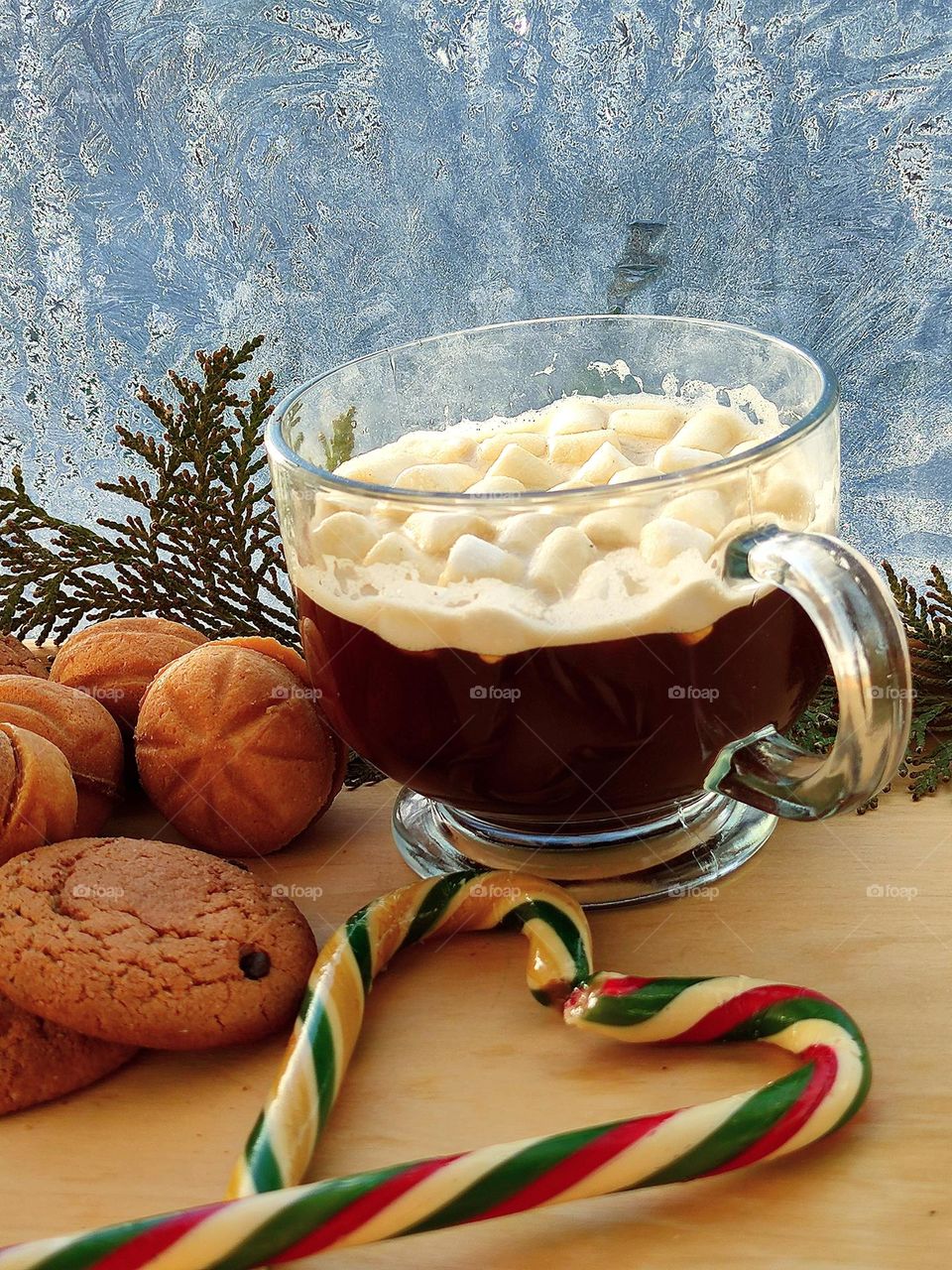 Composition: there is a mug with coffee and marshmallows on the table, next to it there are nut-shaped cookies and oatmeal cookies and two caramel New Year's canes in the shape of a heart. In the background a window with a winter pattern